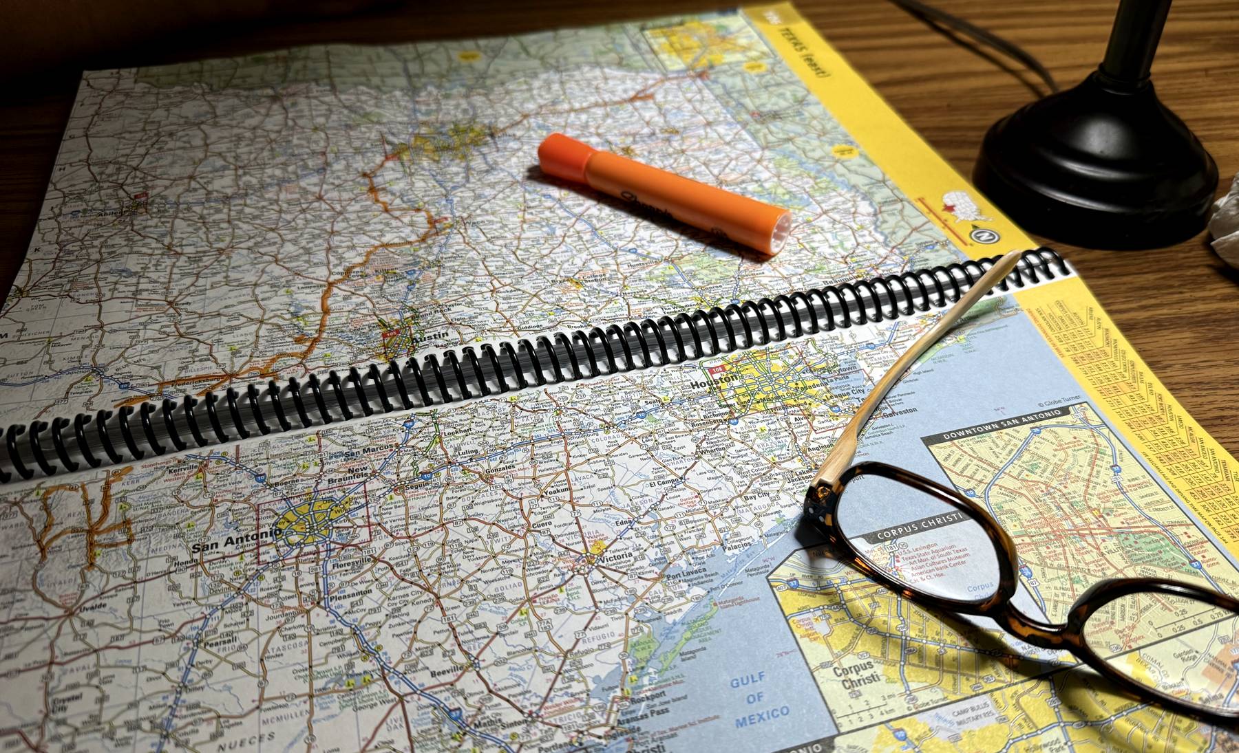 An open spiral-bound road atlas on a desk under lamplight, with an orange highlighter and reading glasses